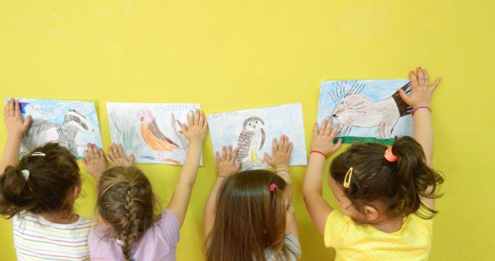 Quattro bimbe di spalle appoggiano i loro disegni sul muro. Un tasso, un pappagallo, una civetta e un istrice.